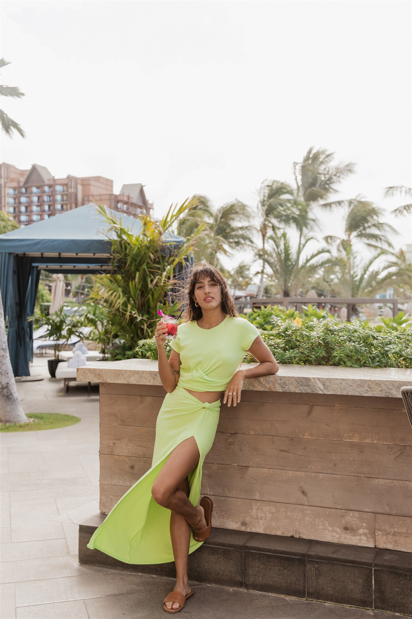 Brunette model facing far front, wearing the Margarita Green Laguna Rib Knot Top Maxi Skirt. This skirt features a ribbed finish, high dramatic slit, and a knot top design. Made from 92% Rayon and 8% Spandex, it's perfect for beach-to-street style. Koy Resort affordable vacation, cruise, and resort-wear.