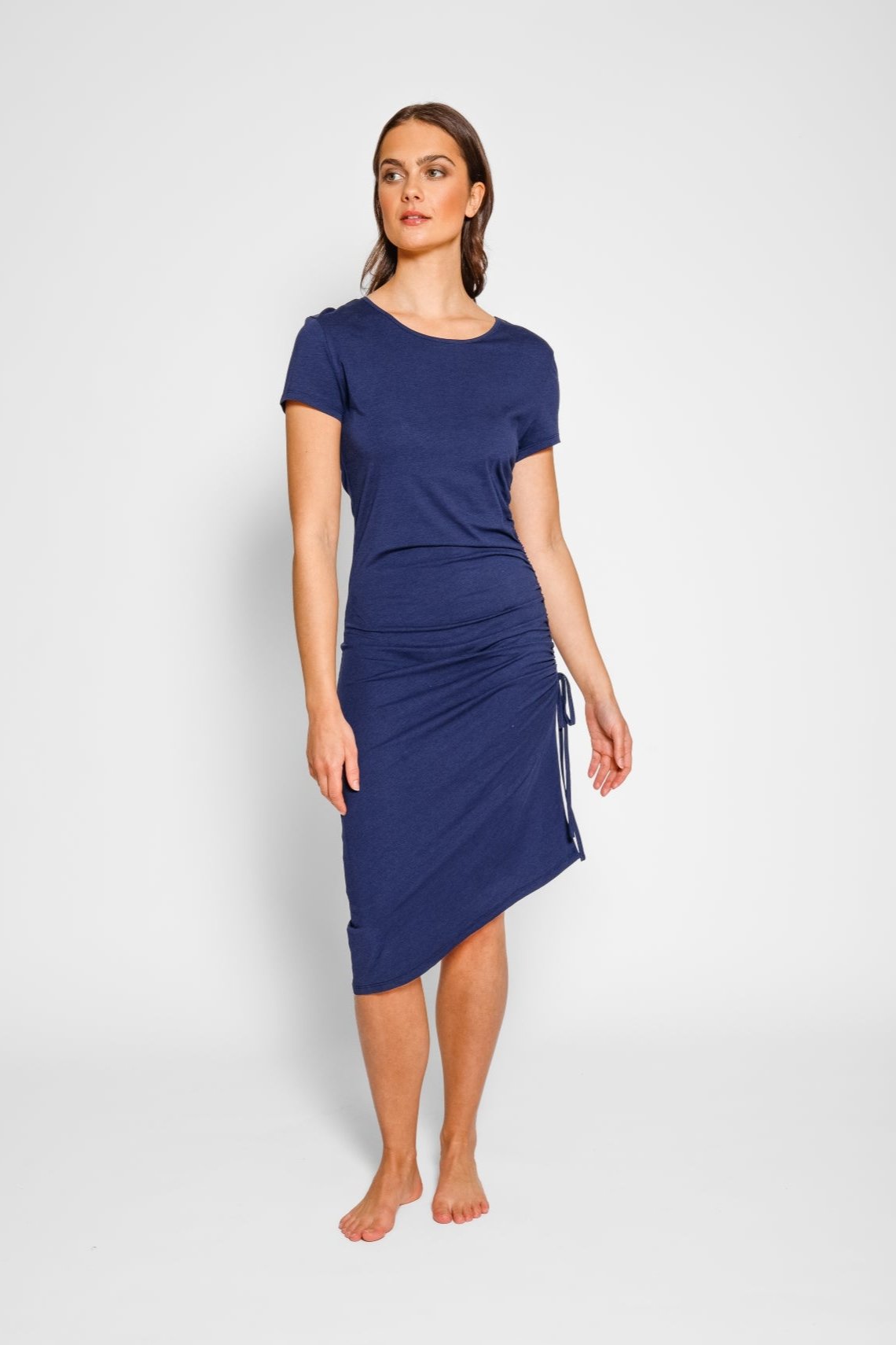 Brunette model facing front, wearing Navy Laguna Beach Midi Dress with adjustable side gathering, scoop neck, short sleeves, asymmetrical hem, and a figure-flattering fit. Made from a cozy cotton jersey blend for stylish and comfortable wear. Koy Resort affordable vacation, cruise, and resort-wear.