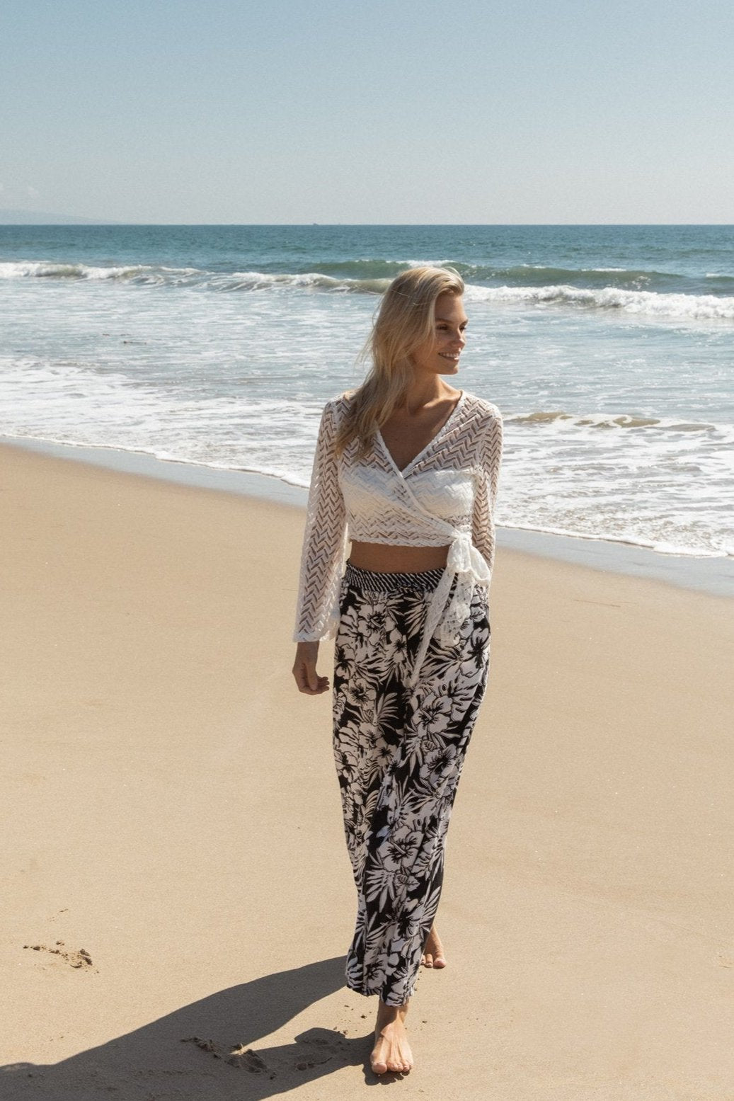 Blonde model facing front, wearing White Zuma Long Sleeve Crochet Wrap Top with sheer crochet herringbone pattern and long bell sleeves. Versatile wrap design available in natural white or black. Perfect for a sassy cover-up look. Koy Resort affordable vacation, cruise, and resort-wear.